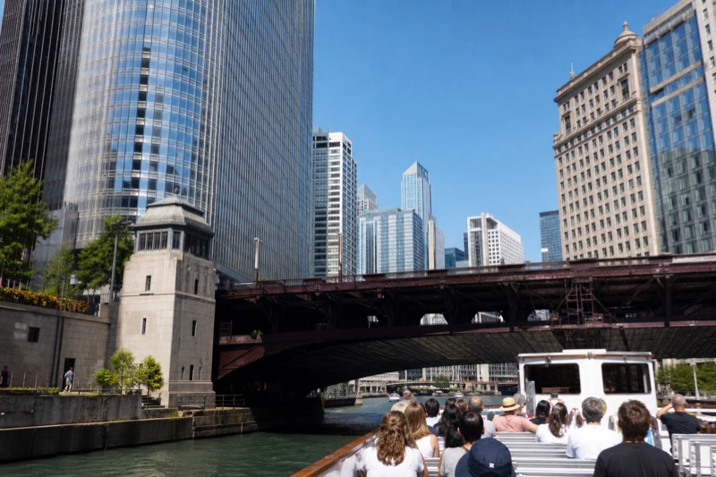 En tursejlads passerer under en lav ståldrejsbro på Chicago River i skarpt middagslys, hvor højhuse rammer udsynet