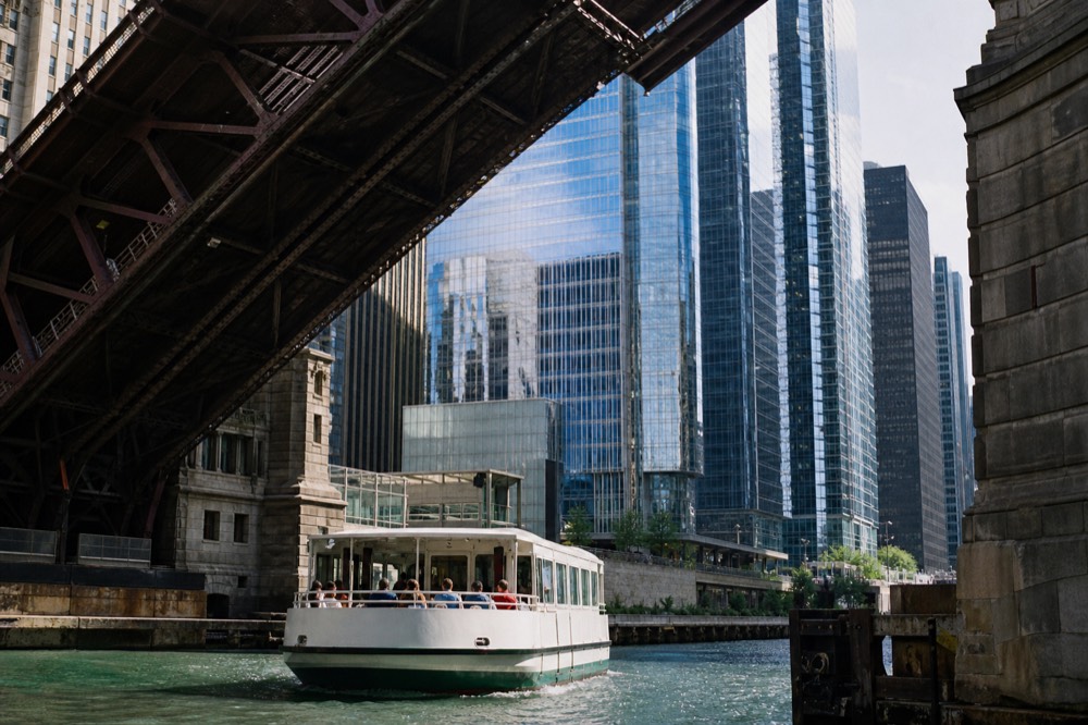 Tour boat passing under a bascule bridge on the Chicago River with skyline towers behind, no text, no logos, no people.